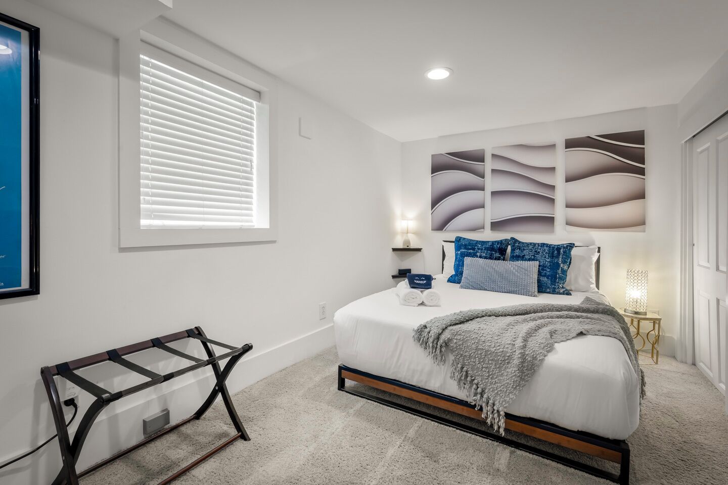 Une chambre à coucher comprenant un lit, une table de chevet, une lampe, une fenêtre avec des stores et une œuvre d’art accrochée au mur.