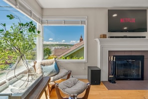 Ein Wohnzimmer mit Fernseher, Kamin und Blick auf eine Stadtlandschaft durch ein großes Fenster.
