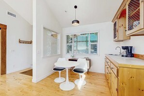 A modern kitchen with a wooden countertop, a dining area with two white chairs, and a large window with blinds. - Pinetree Palace | Mountain Retreat | Games | Treetop Views | Sleeps 6 (Lake Arrowhead)