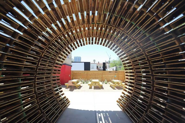 Une structure arquĂ©e en bois, dotĂ©e de bancs et de tables, qui mĂšne Ă un espace extĂ©rieur oĂč lâon aperçoit des bĂątiments en arriĂšre-plan.