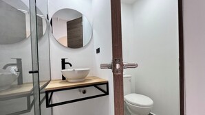 Un baño moderno con cabina de ducha de vidrio, un lavamanos blanco con estante de madera, un espejo redondo y un inodoro blanco.
