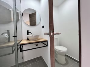 Un bany modern amb plat de dutxa de vidre, un lavabo blanc amb prestatge de fusta, un mirall rodó i un vàter blanc.