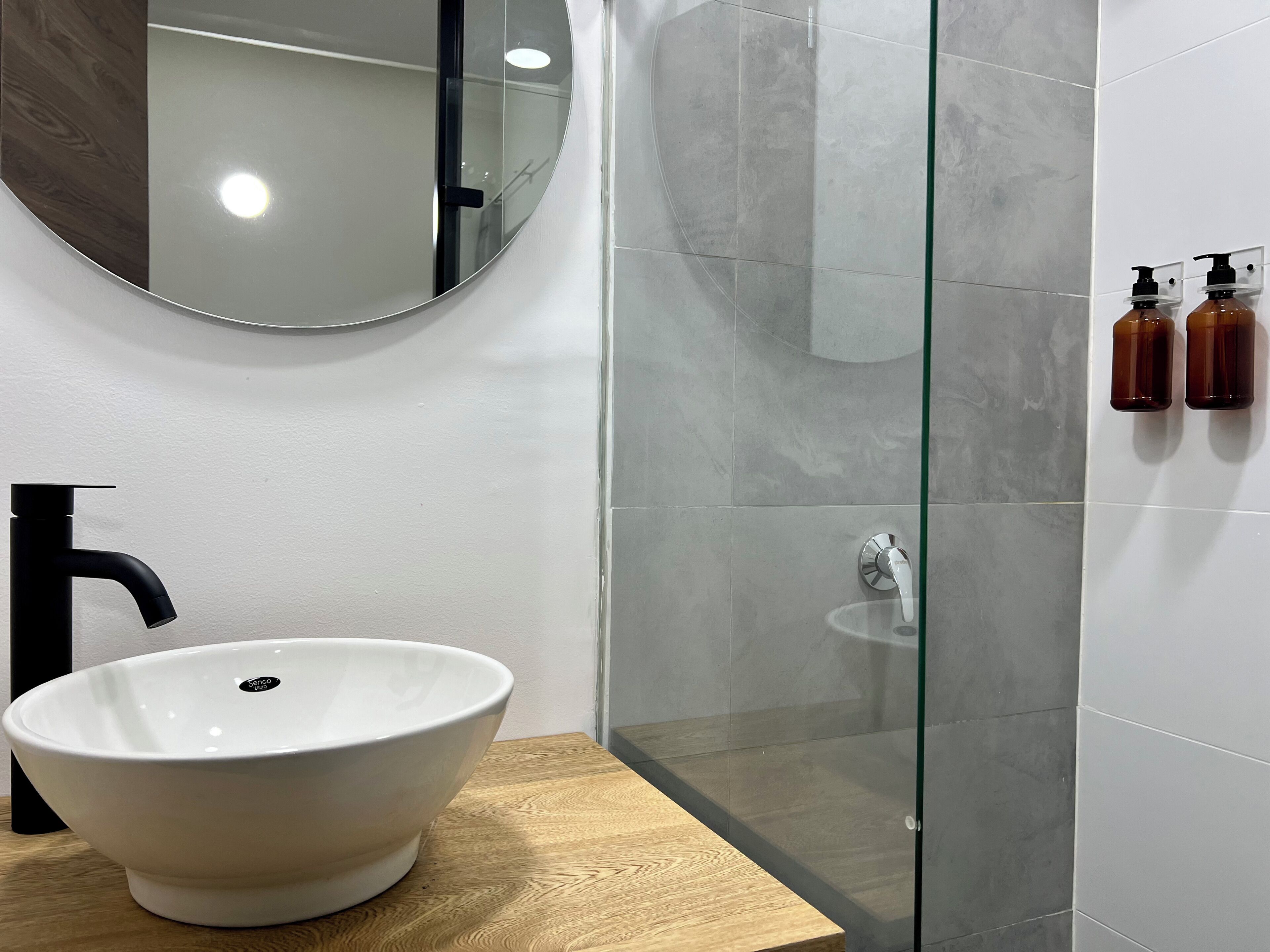 Une salle de bain moderne avec un grand lavabo blanc sur pied, un robinet noir, un miroir rond, une cabine de douche en verre et deux distributeurs de savon fixés au mur.