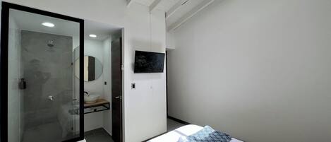 Een moderne hotelkamer met een glazen douchecabine, een wandgemonteerde televisie en een bed met een gestreept kussen.