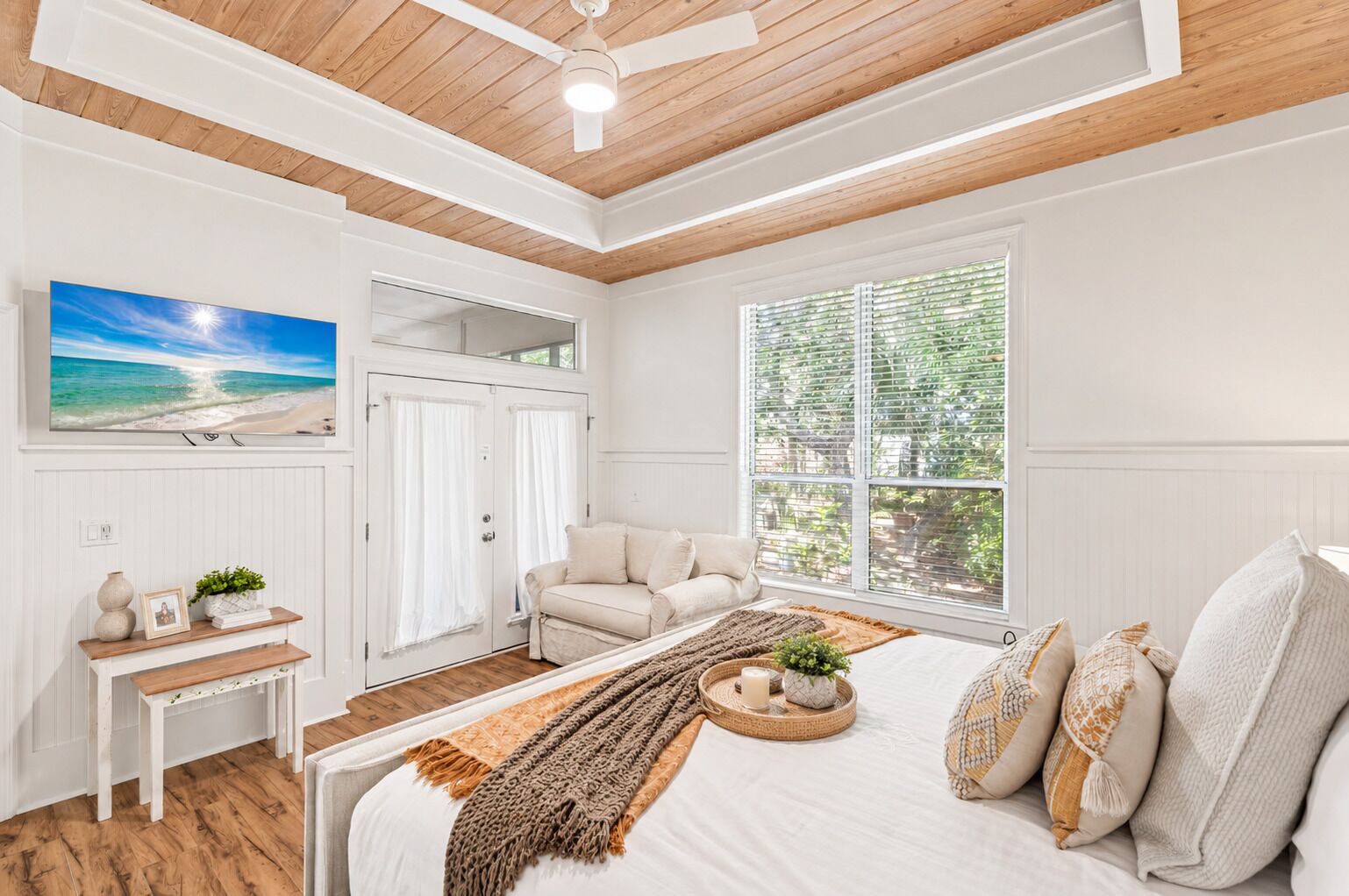 Ein Schlafzimmer mit Bett, Deckenventilator, Fenster mit Jalousien, einer Tür und einem an der Wand montierten Fernseher.