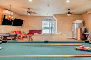 Une table de billard avec des queues de billard, un canapé, un téléviseur et des ventilateurs de plafond dans une pièce aux murs de couleur beige.