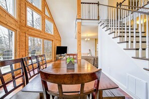 A dining area with a wooden table and chairs, a TV, and a staircase with a railing. - Lake Anna A-Frame | Beach, Dock & Hot Tub | 5BR (Bumpass)