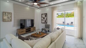 A modern living room with a flat-screen TV, a ceiling fan, a sofa, a coffee table, and a view of a swimming pool. - Casa De La Puerta Azul (San Felipe Puerto Plata)