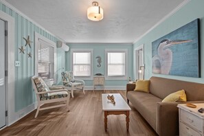 A room with a wooden floor, a white coffee table, a brown sofa, a white armchair, and a painting of a bird on the wall. - The Lookout at Sandy Sea Star: 2 Bdrm Upper Unit Near Beach Access (Carolina Beach)