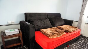 Ein Zimmer mit einer schwarzen Couch, einem roten Kissen, einem Holztischchen mit einem gefalteten Handtuch und einem gemusterten Teppich.