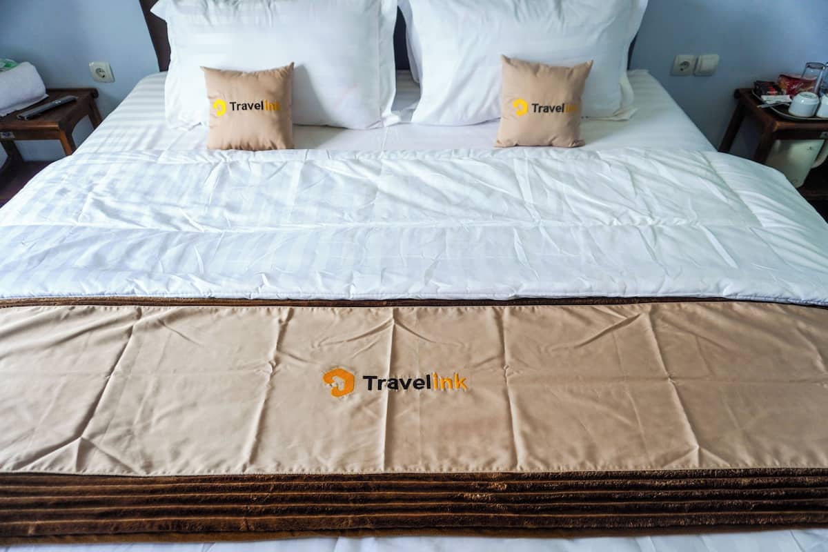 Una cama con ropa de cama blanca, dos almohadas con el logotipo de Travelink y un edredón marrón.