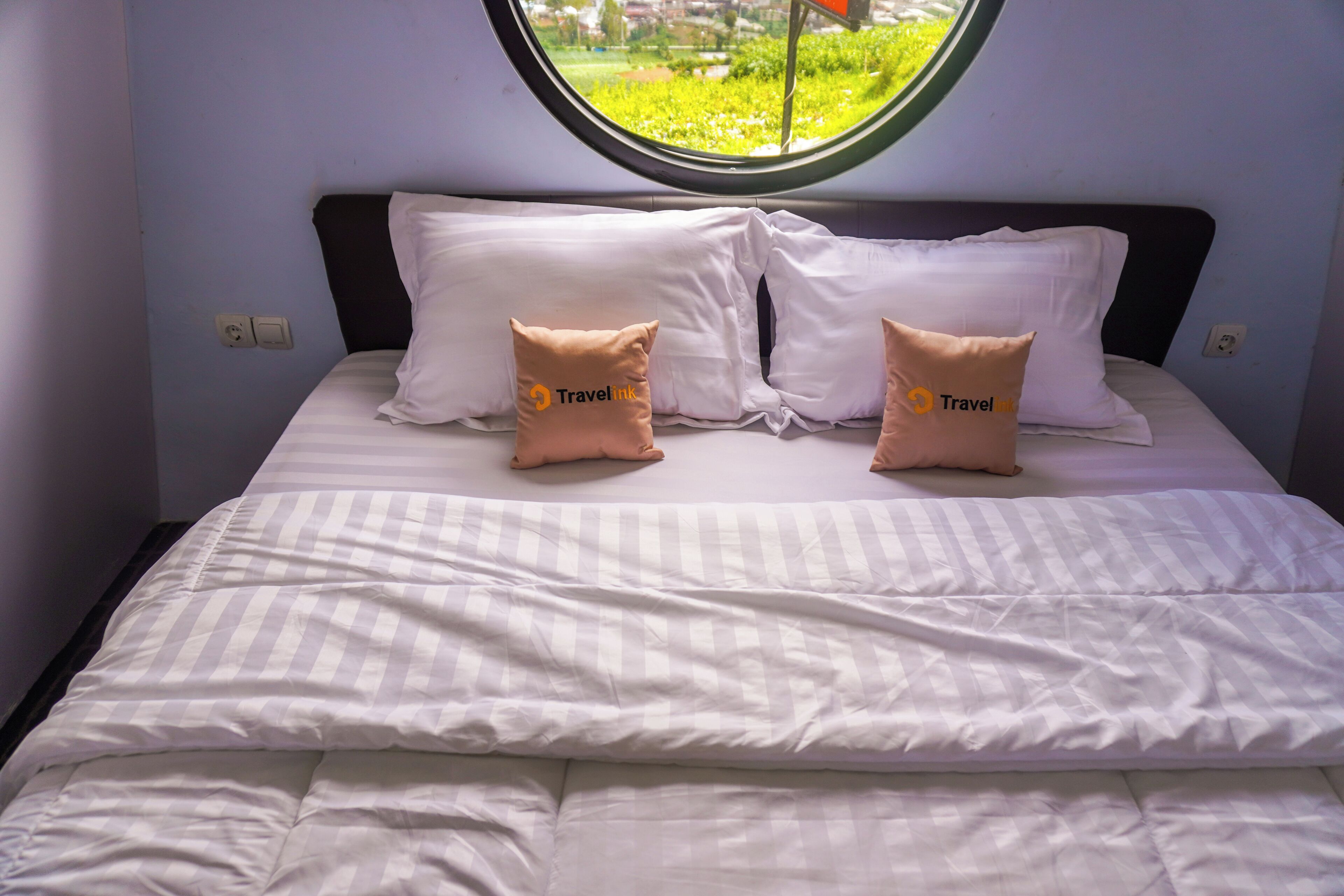 Una cama con ropa blanca y dos almohadas con la marca 'Travelink', una ventana que da a zonas verdes y un enchufe de pared.