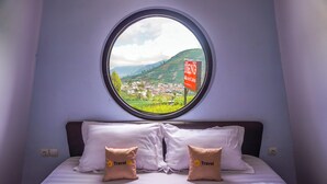 Ein Bett mit zwei Kissen, ein rundes Fenster mit Blick auf eine Stadt und Hügel sowie ein Schild mit der Aufschrift 'DIENG'.