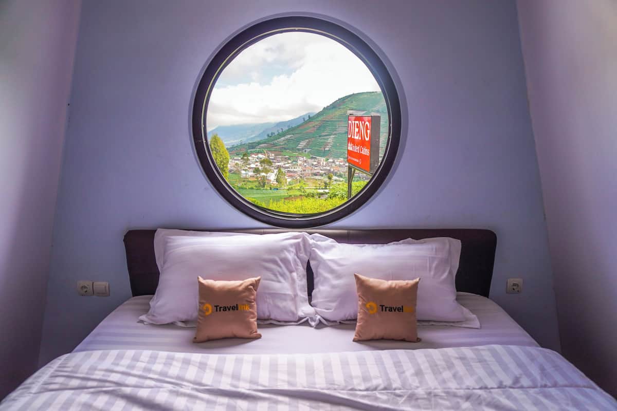 Una cama con dos almohadas, un ventanal redondo con vistas a un pueblo y colinas, y un cartel que dice 'DIENG'.