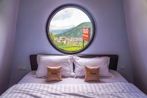 Un lit avec deux oreillers, une fenêtre ronde donnant sur une ville et des collines, et une pancarte sur laquelle on peut lire « DIENG ».