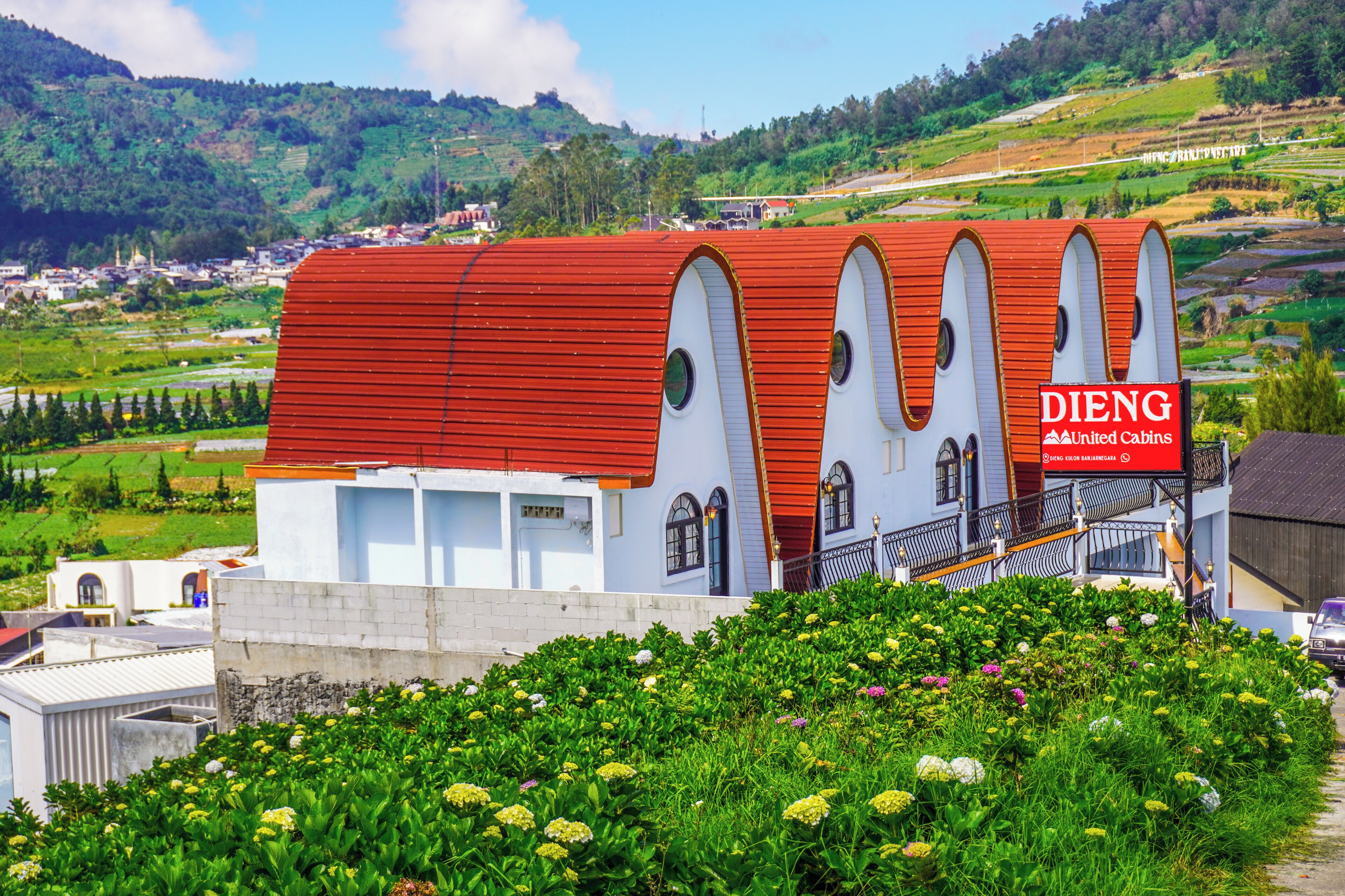 Un bâtiment avec un toit rouge et des murs blancs, une enseigne indiquant « DIENG United Cabins », et un jardin luxuriant au premier plan.