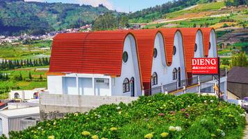 A building with a red roof and white walls, a sign for 'DIENG United Cabins,' and a lush garden in the foreground.