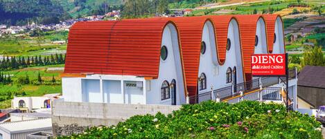 Un bâtiment avec un toit rouge et des murs blancs, une enseigne indiquant « DIENG United Cabins », et un jardin luxuriant au premier plan.