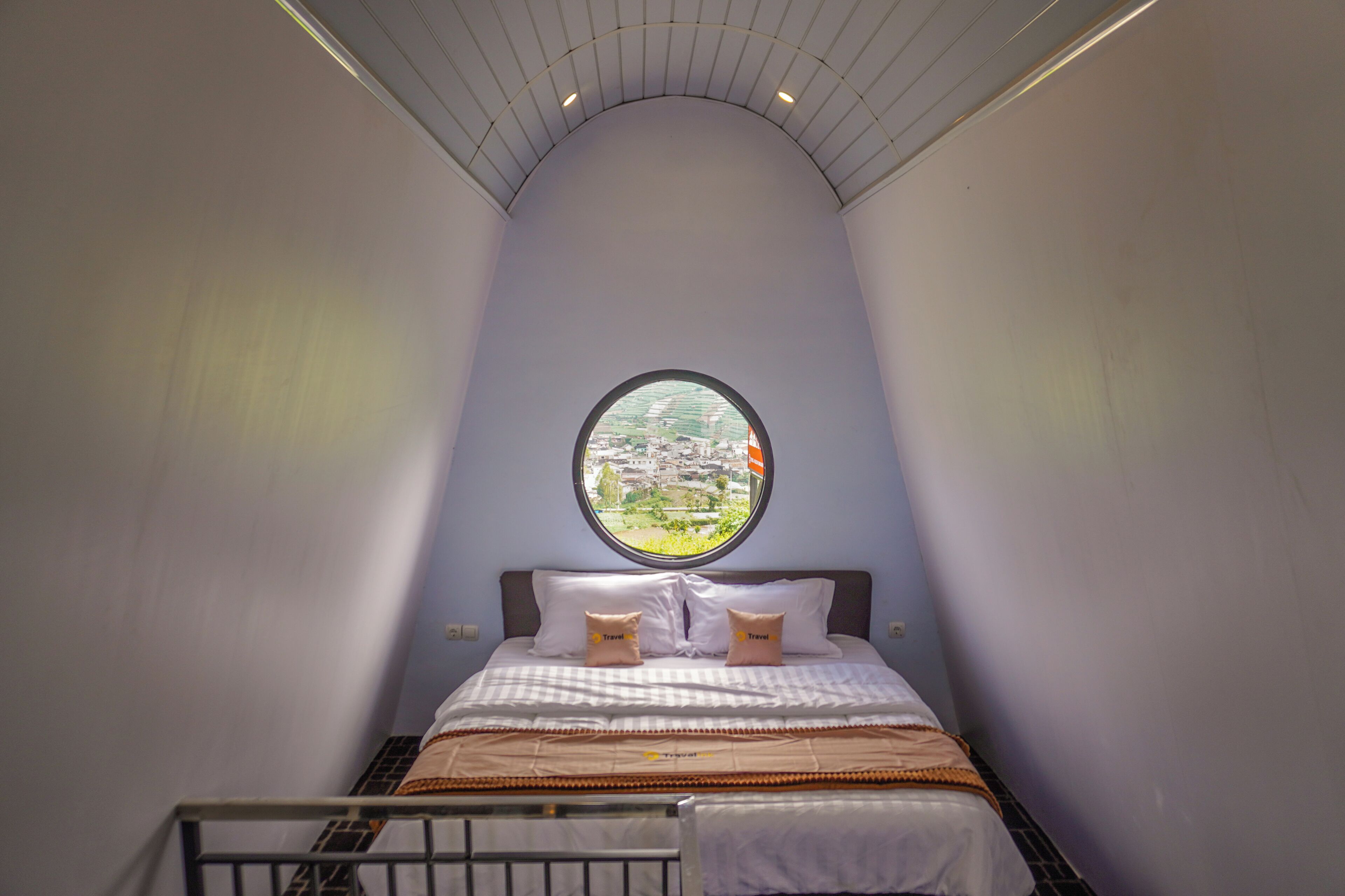 A bedroom with a curved ceiling, a round window, a bed with a checkered bedspread, and two pillows labeled 'Travel.'