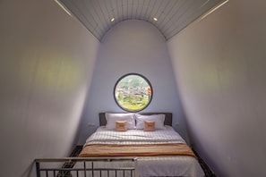 Een slaapkamer met een gebogen plafond, een rond raam, een bed met een geruit sprei en twee kussens met de tekst 'Travel'.