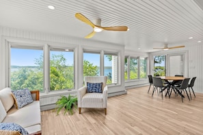 A spacious room with large windows, a ceiling fan, a dining table with chairs, and a cushioned seating area. - Plumb Island Sound Waterfront Sunset Getaway (Ipswich)