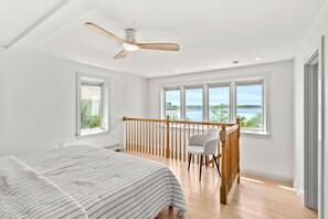 A bedroom with a bed, a chair, a balcony with a railing, and large windows. - Plumb Island Sound Waterfront Sunset Getaway (Ipswich)
