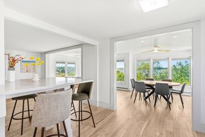 A modern kitchen with a white countertop, bar stools, and a dining area with a wooden table and chairs. - Plumb Island Sound Waterfront Sunset Getaway (Ipswich)