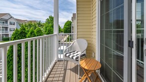 A balcony with a white railing, a wicker chair with a striped cushion, and a small wooden table. - End of the Season Special! Beautiful Rehoboth Beach Condo Pools Tennis Parking (Rehoboth Beach)