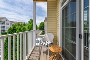 A balcony with a white railing, a wicker chair with a striped cushion, and a small wooden table. - Beautiful Rehoboth Beach Condo Pools Tennis Parking (Rehoboth Beach)