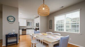 A dining area with a wooden table, white chairs, a woven pendant light, and a kitchen in the background. - End of the Season Special! Beautiful Rehoboth Beach Condo Pools Tennis Parking (Rehoboth Beach)