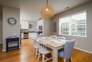 A dining area with a wooden table, white chairs, a woven pendant light, and a kitchen in the background. - Beautiful Rehoboth Beach Condo Pools Tennis Parking (Rehoboth Beach)