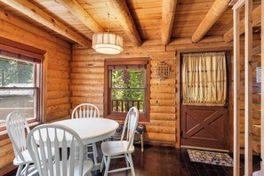 Interior de una cabaña de troncos con una mesa de comedor, sillas, una lámpara colgante, una ventana con vistas y una puerta con cortinas.