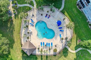 Vista aérea de una piscina circular con sillas de descanso, palmeras y un edificio pequeño.