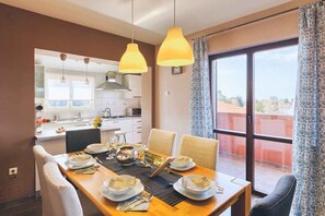 A dining area with a table set for four, a kitchen in the background, and a large window with curtains. - Holiday apartment with sea view (Banjole)