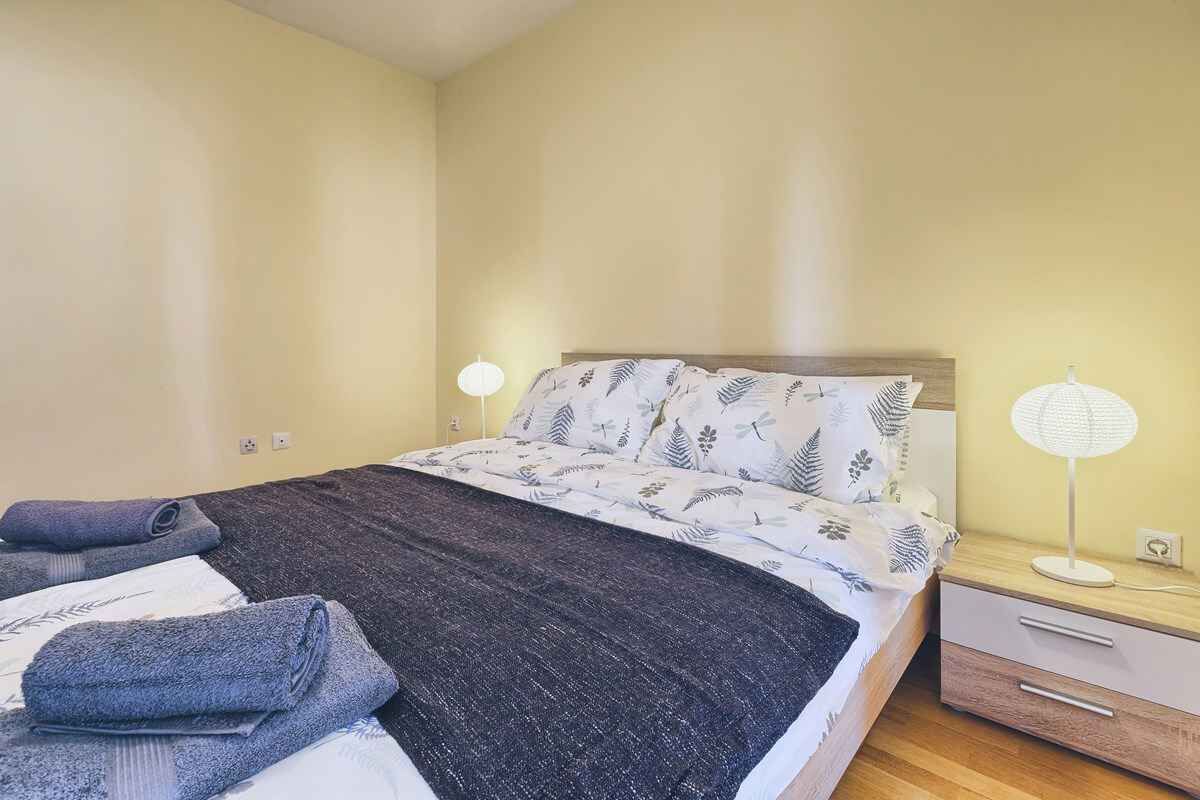Un lit bien fait, avec une couvre-lit à motifs, deux lampes de chevet, une petite commode et une prise électrique au mur.