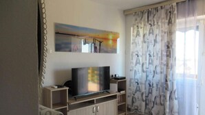 Uma sala de estar com televisão de tela plana sobre um suporte de madeira, um quadro acima da TV e uma janela com cortinas estampadas.