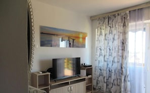A living room with a flat-screen TV on a wooden stand, a painting above the TV, and a window with patterned curtains. - Holiday home with pool and air conditioning (Csopak)