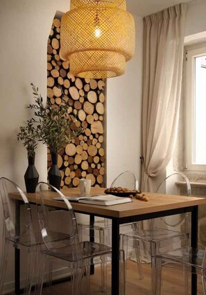 Ein Essbereich mit Holztisch, klaren Stühlen, einer gewobenen Leuchte, einer Vase mit Grünpflanzen und einer Holzwand.