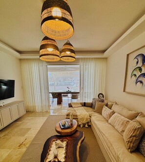 A living room with a sofa, a coffee table, a TV, and a large window with curtains. - Relaxing Beach Apartment with pool, jacuzzi, beach in RivieraGolf Playa Dorada (Playa dorada)
