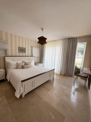 A bedroom with a large bed, a bedside table, a hanging light fixture, and a window with curtains. - Relaxing Beach Apartment with pool, jacuzzi, beach in RivieraGolf Playa Dorada (Playa dorada)