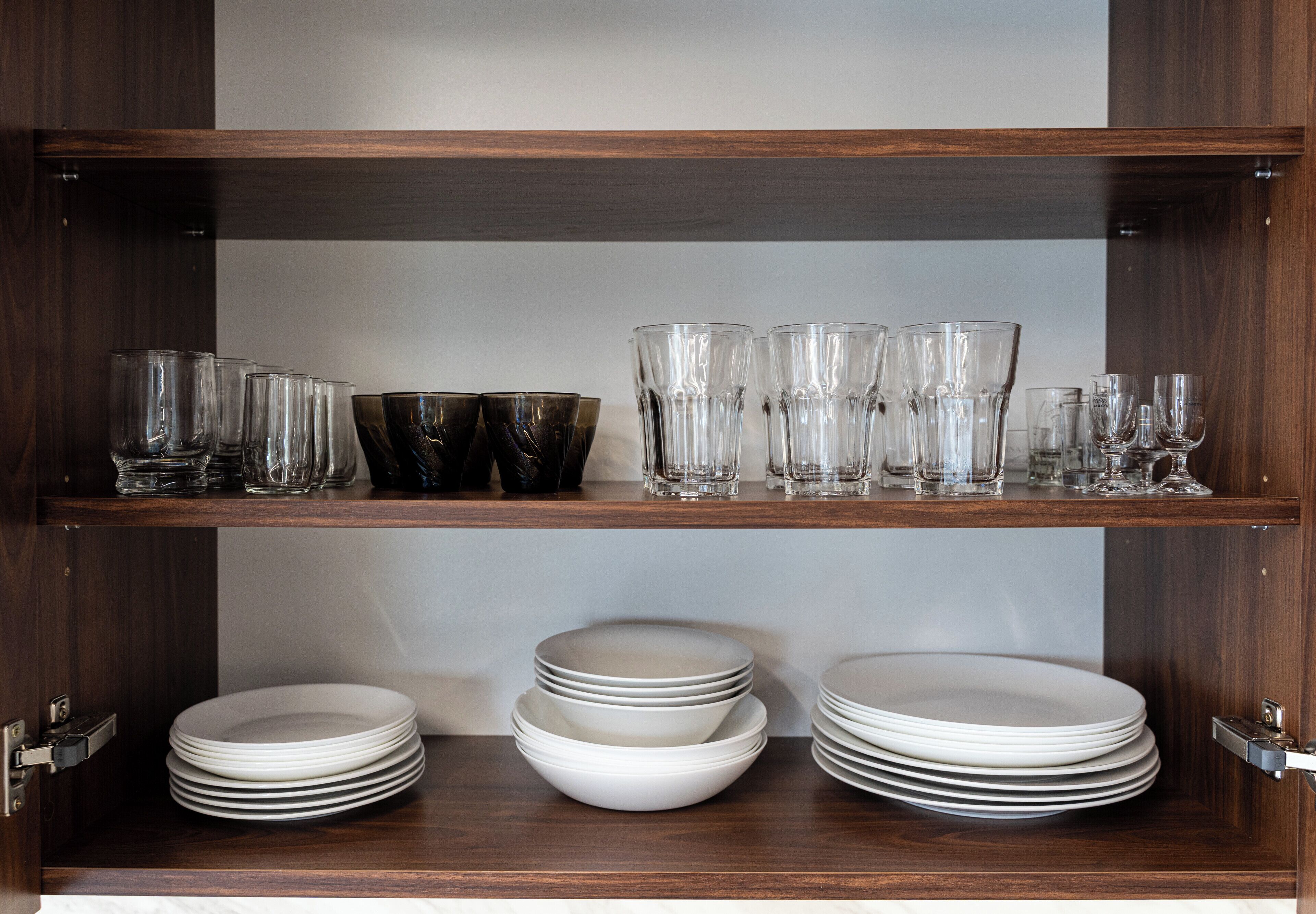 A wooden cabinet with shelves holding clear glasses, black bowls, and white plates.