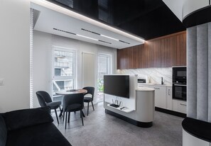 Et moderne køkken med indbygget ovn, et spiseområde med et rundt bord og stole, samt et TV ophængt på et hvidt skab.