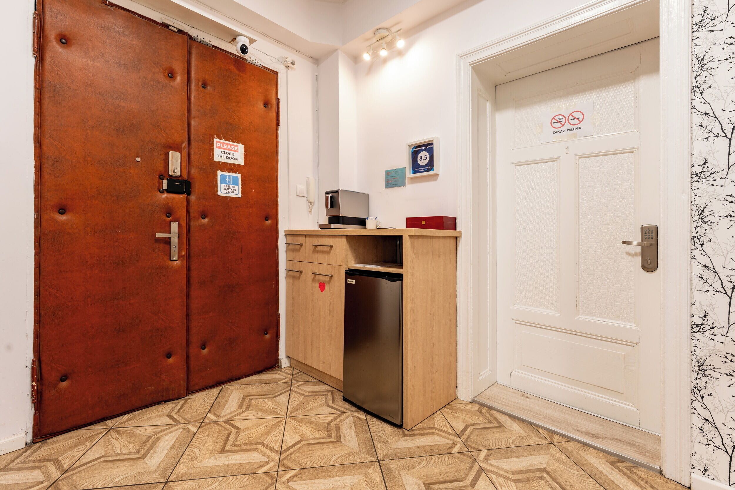 A room with a wooden wardrobe, a small wooden cabinet with a refrigerator, a microwave, and a red box on top. There are two doors, one with a 'Please close the door' sign and another with a 'No smoking' sign.