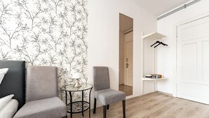 Um quarto com duas cadeiras cinzentas, uma mesa redonda pequena com abajur, uma porta branca e um cabideiro.