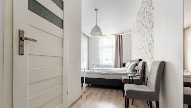 Quarto com cama, cadeira, mesa de cabeceira, luminária pendente e janela com cortinas.