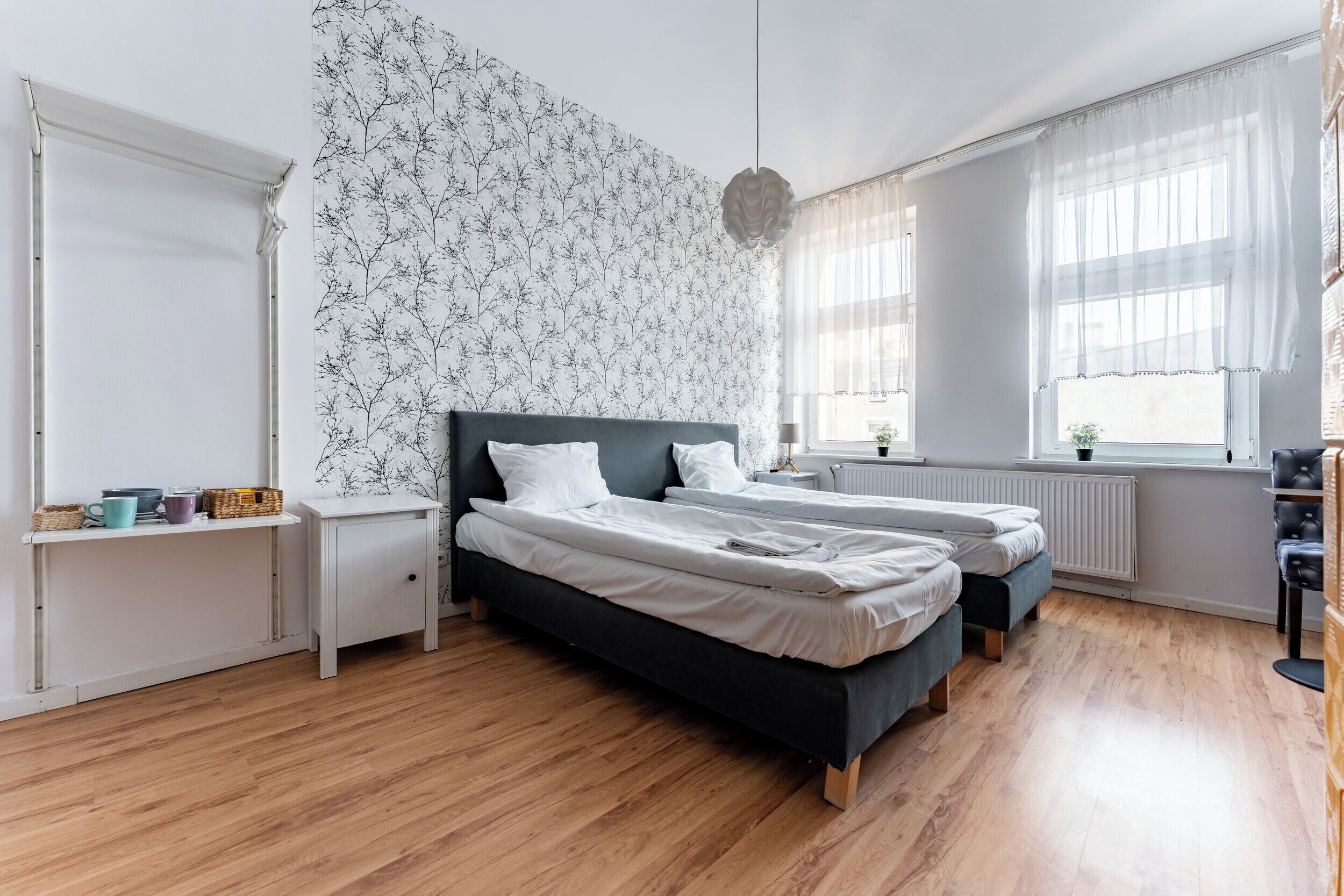A modern bedroom with two beds, a white wall with a tree pattern, wooden flooring, and a window with sheer curtains.