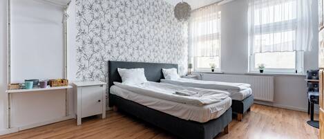 A modern bedroom with two beds, a white wall with a tree pattern, wooden flooring, and a window with sheer curtains.