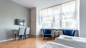 Ett hotellrum med trägolv, en säng, ett skrivbord med en stol, en tv och ett fönster med gardiner.
