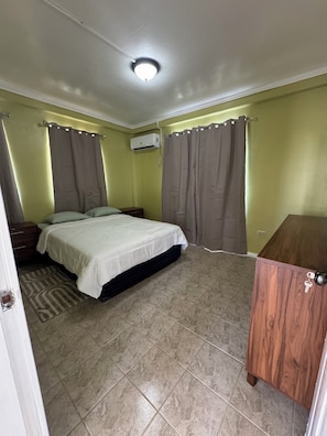 Un dormitorio con una cama, cortinas, un equipo de aire acondicionado, un armario de madera y una alfombra estampada.