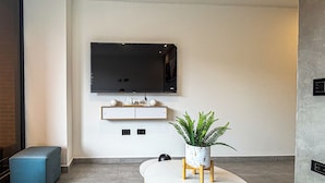 Una sala de estar moderna con un televisor de pantalla plana montado en la pared, una pequeña consola blanca, un helecho en maceta y un reposapiés azul.